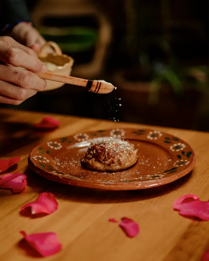 Fotografía de Na Nena: una mano espolvorea queso sobre una garnacha istmeña servida en un plato de barro con pétalos de rosa